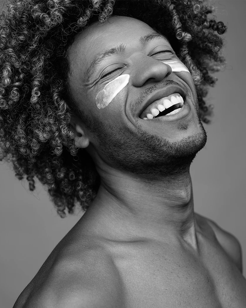 Black and white close-up of a smiling man with curly hair, showing his joy and confidence. He has two streaks of skincare product under his eyes, highlighting a focus on skincare and self-care. The image emphasises the effectiveness of skincare treatments, reflecting healthy, well-cared-for skin.