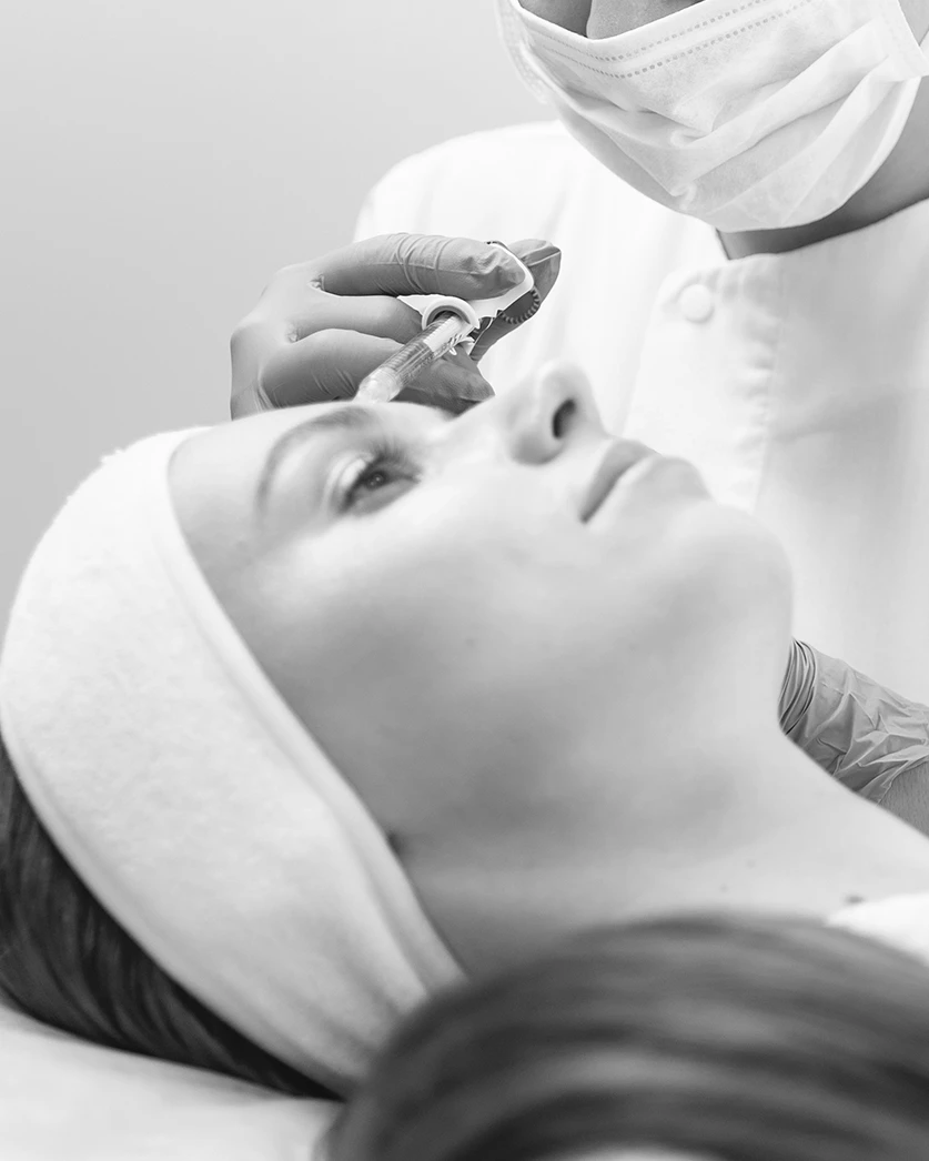 Black and white image of a woman receiving a facial treatment. She is lying down with a calm, content expression while a practitioner wearing black gloves prepares to apply a facial mask using a brush. The setting exudes a serene and professional atmosphere, highlighting the quality and care provided at Beyond Aesthetix Clinic.