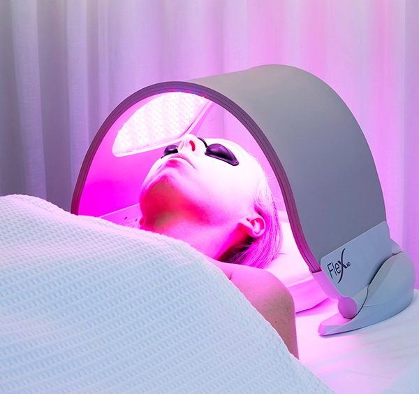 Individual undergoing Dermalux LED Light Therapy, lying comfortably with protective eyewear under a specialized LED light device emitting pink light. The therapy targets the skin to stimulate collagen production, reduce inflammation, and promote overall skin rejuvenation. The serene setting with soft, ambient lighting ensures a relaxing and effective treatment experience.
