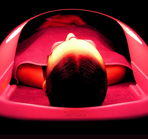 Person undergoing Dermalux LED Light Therapy, lying under a specialized LED light device. The red light targets the skin to activate cellular activity, promoting collagen production and enhancing skin rejuvenation. The individual is covered with a towel, creating a relaxing and soothing environment.