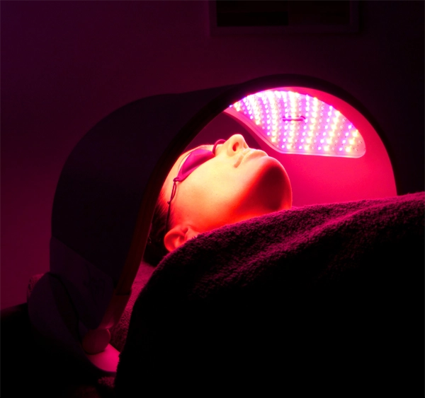 Individual receiving Dermalux LED Light Therapy under a specialized LED device emitting red light. The therapy targets the skin to stimulate collagen production, reduce fine lines, and promote skin rejuvenation. The individual is lying comfortably with protective eyewear, creating a serene and effective treatment environment.